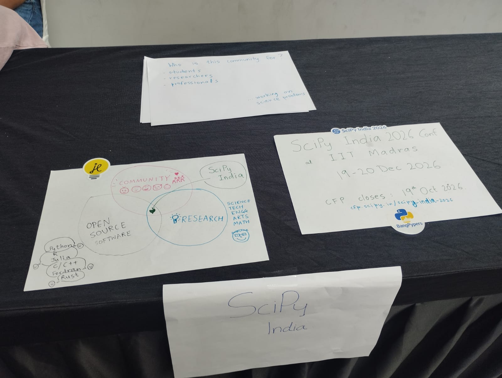 Hand-drawn signs on the booth table about the community and the SciPy India 2026 conference