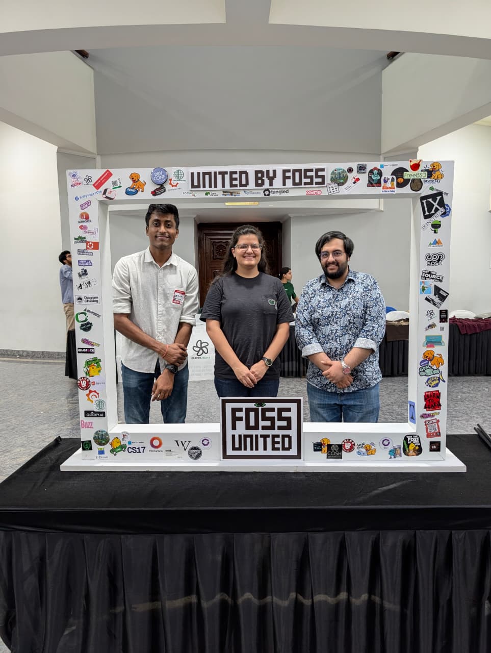 Three people posing inside the "United by FOSS" photo frame at IndiaFOSS 2026